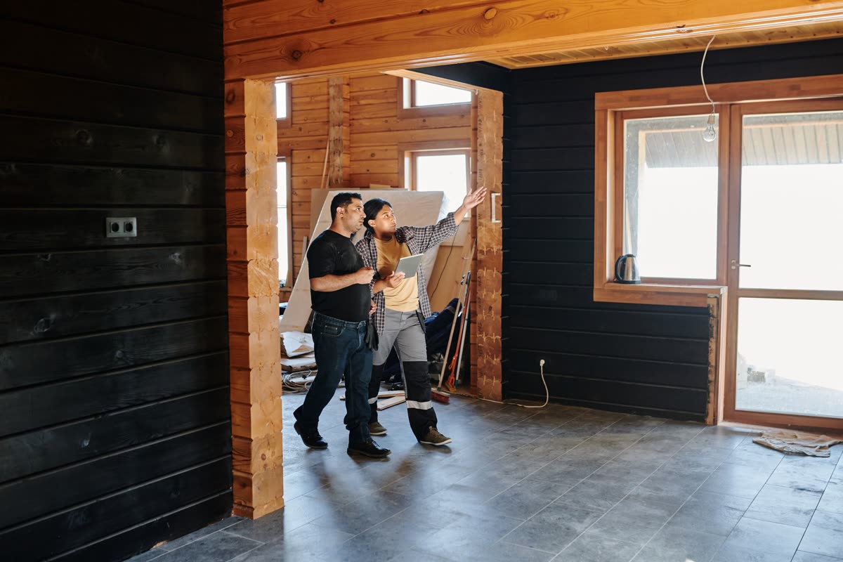 A renovation professional with a tablet walks a homeowner through planned work in a bright, wood-panelled home.